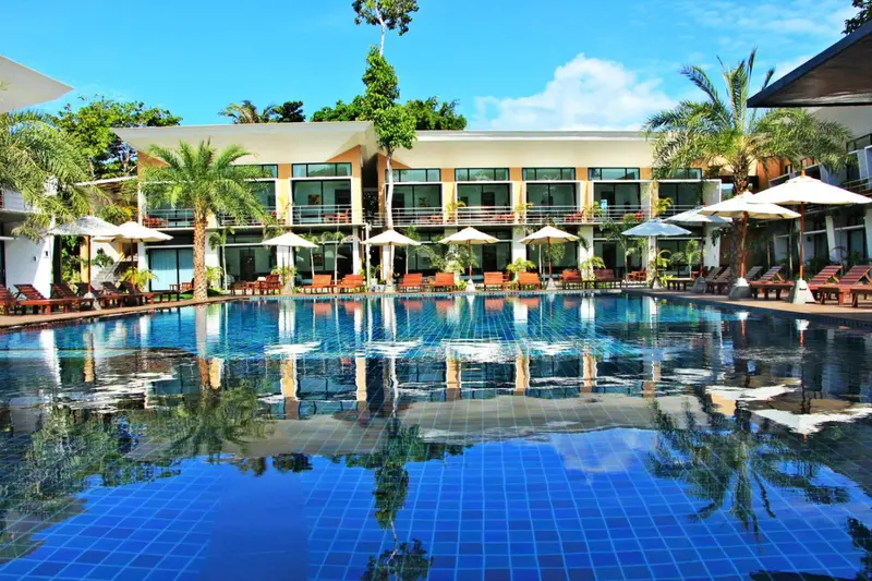 Une piscine turquoise reflète un bâtiment blanc entouré de végétation tropicale et de transats.