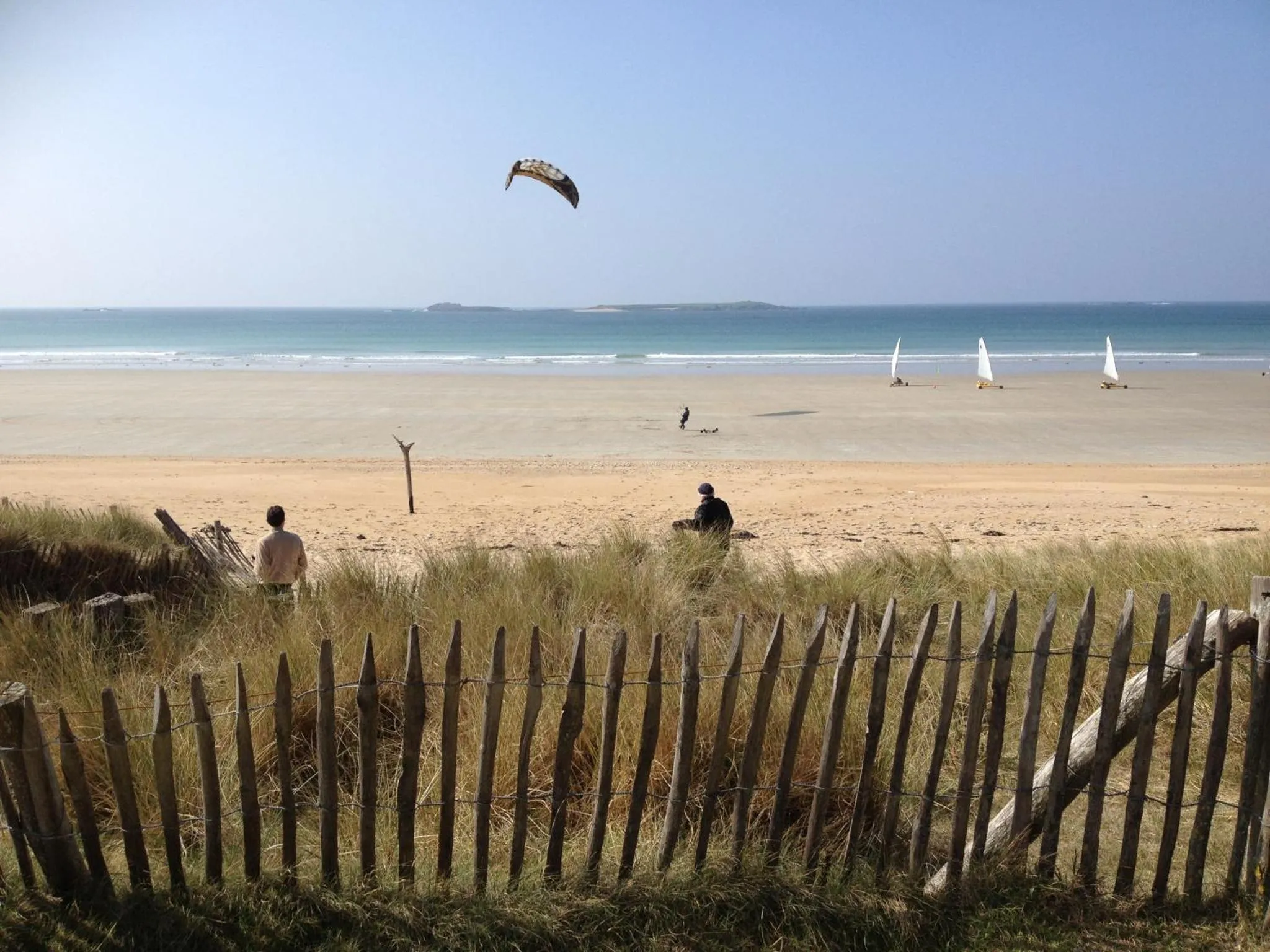Une plage de sable fin avec des personnes assises et des voiles dans le ciel.
