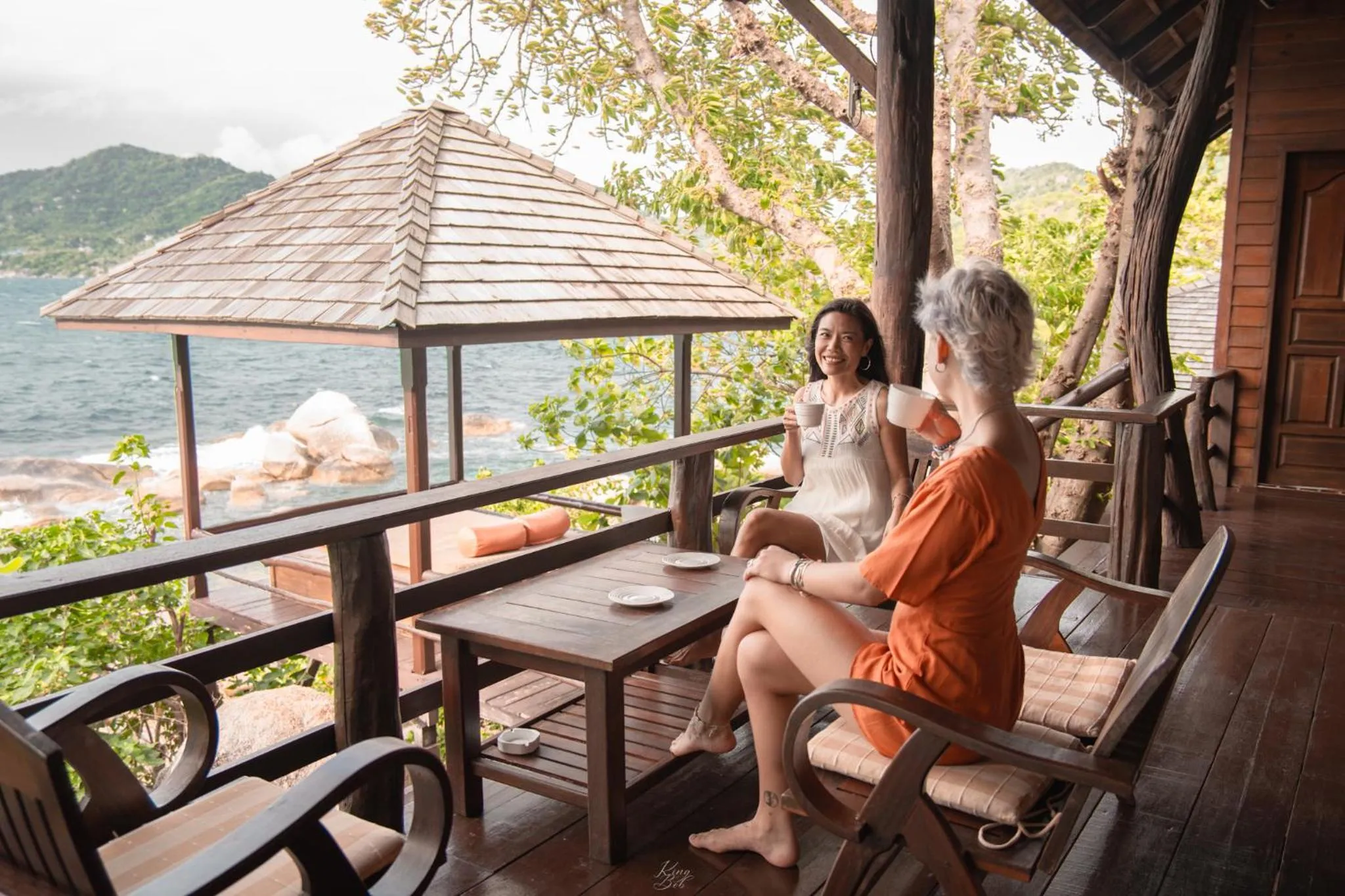 Deux femmes discutent autour d'une table sur une terrasse en bois avec vue sur la mer.