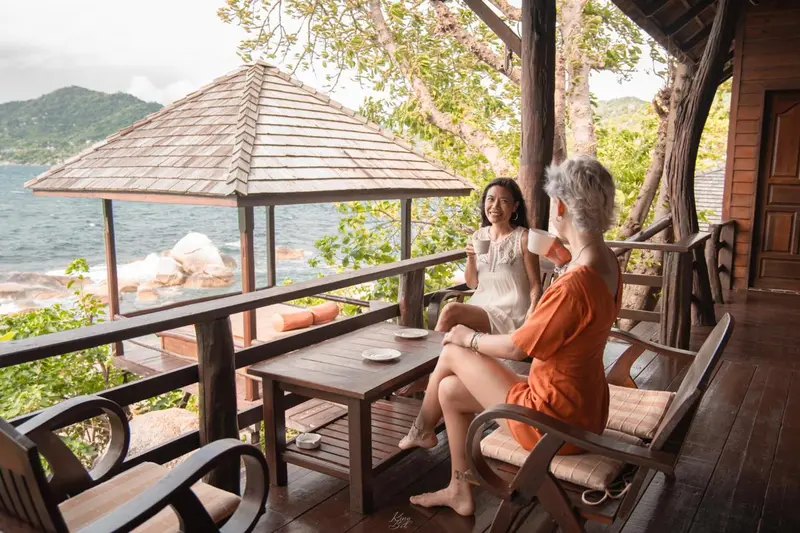 Deux femmes discutent autour d'une table sur une terrasse en bois avec vue sur la mer.
