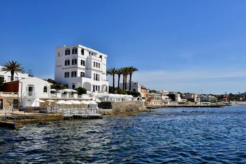 Bâtiment blanc à plusieurs étages surplombant une côte rocheuse et une mer bleue.