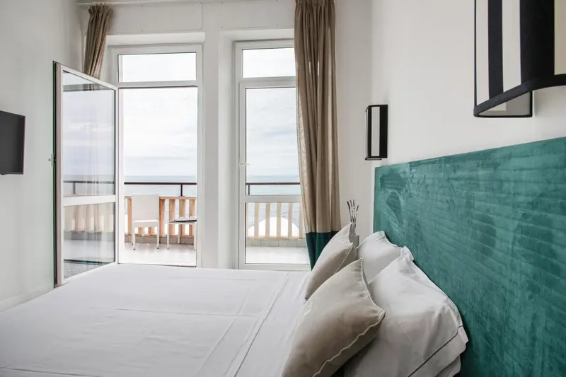 Une chambre lumineuse avec un lit blanc et un accès à un balcon avec vue sur la mer.