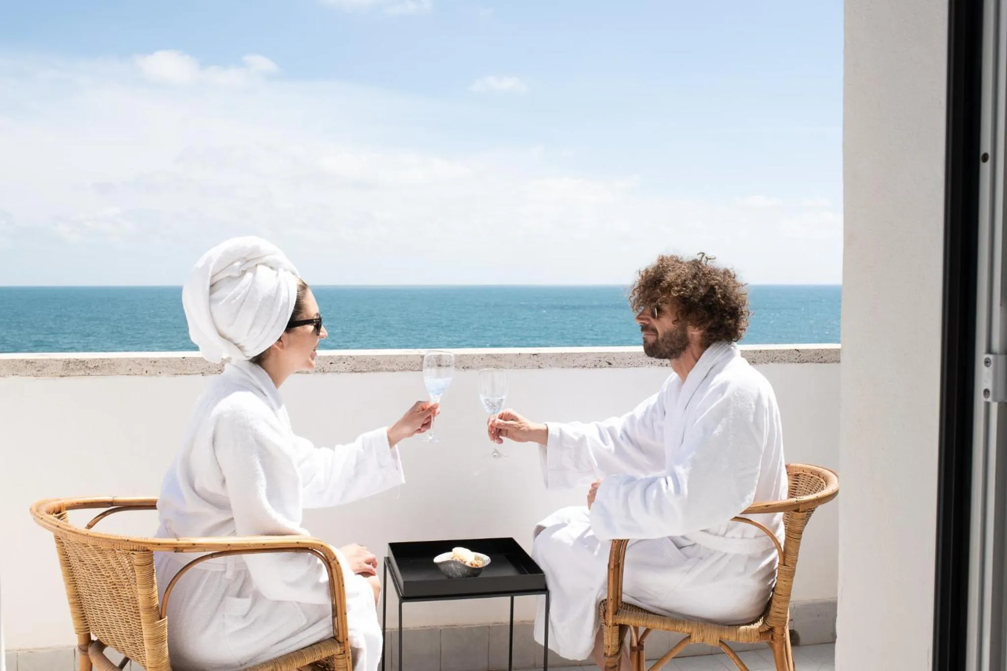 Un couple en peignoir trinque sur une terrasse avec vue sur la mer.