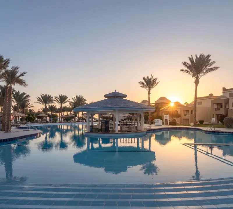 Une piscine rectangulaire reflète le ciel orangé du coucher de soleil et un kiosque à colonnes.