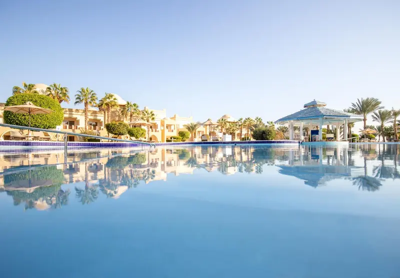Vue d'une piscine turquoise reflétant un bâtiment blanc et des palmiers sous un ciel bleu.