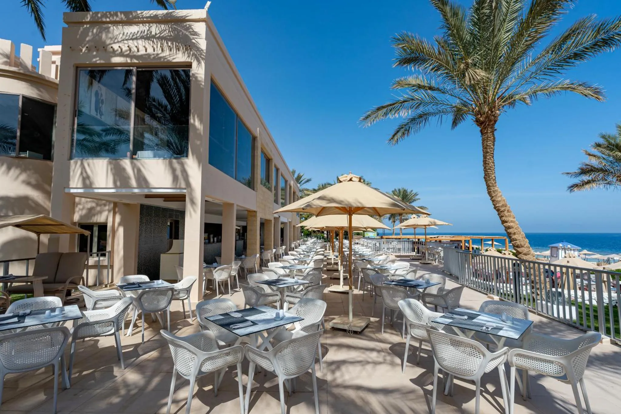 Terrasse ombragée avec tables et chaises en plastique blanc, vue sur la mer.