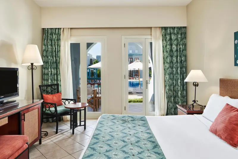 Chambre lumineuse avec un lit et un coin salon donnant sur une piscine.