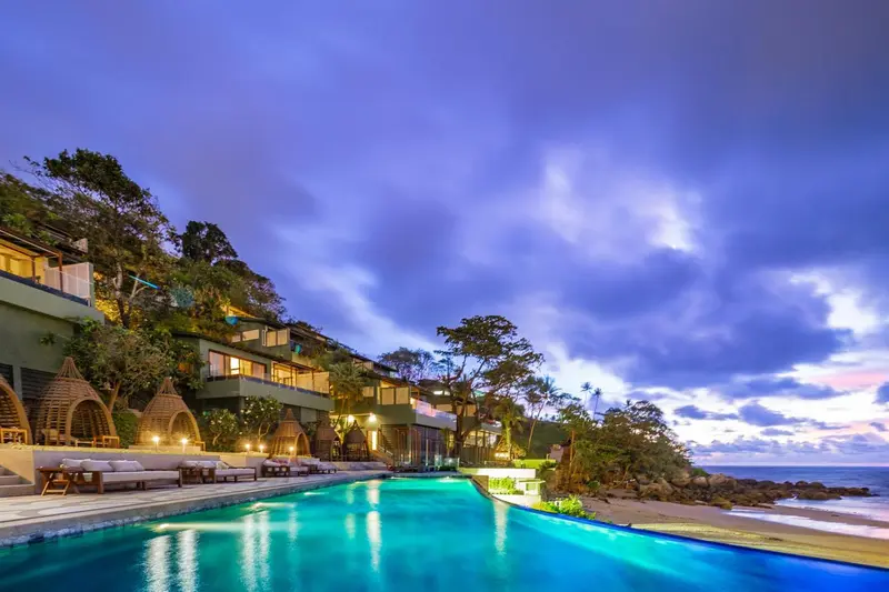 Une piscine à débordement lumineuse surplombe une plage au coucher du soleil.