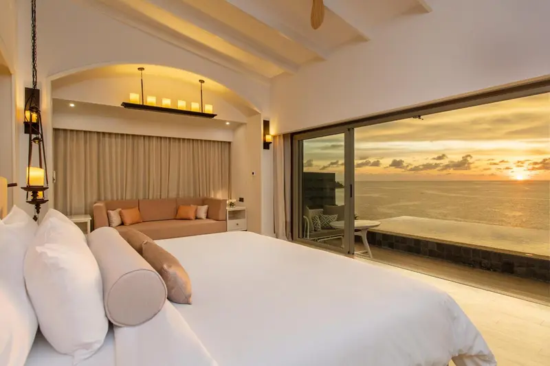 Une chambre lumineuse avec un grand lit donnant sur une vue mer au coucher du soleil.