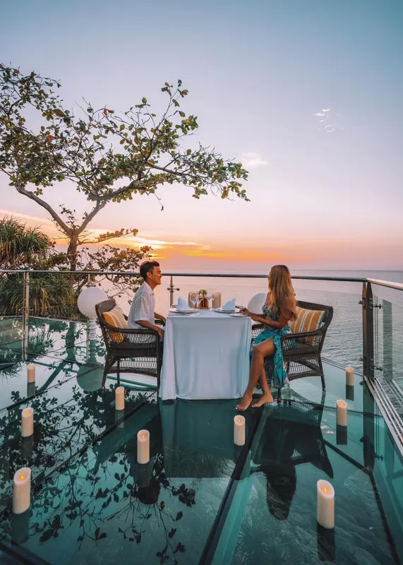 Un couple dîne à une table dressée sur une terrasse vitrée surplombant la mer au coucher du soleil.