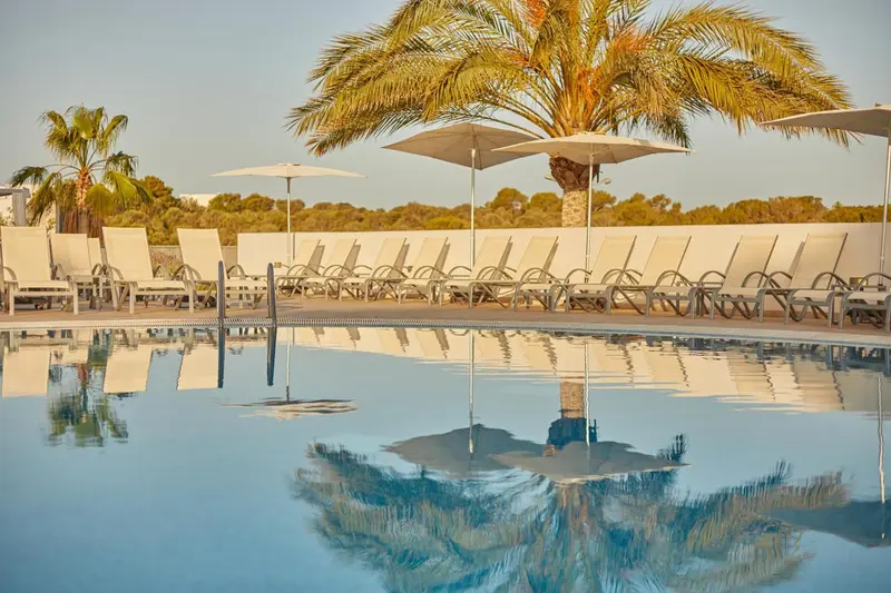 Transats blancs alignés au bord d'une piscine reflétant le ciel et les palmiers.