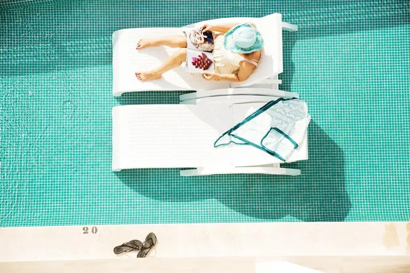 Deux transats blancs au bord d'une piscine turquoise, avec des personnes se relaxant et lisant.