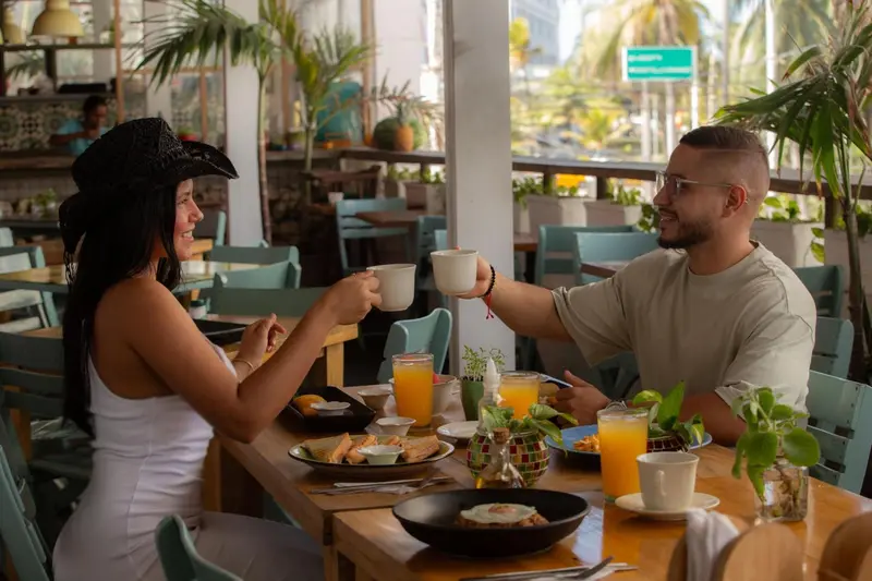 Deux personnes sourient en levant leurs tasses dans un restaurant ensoleillé.
