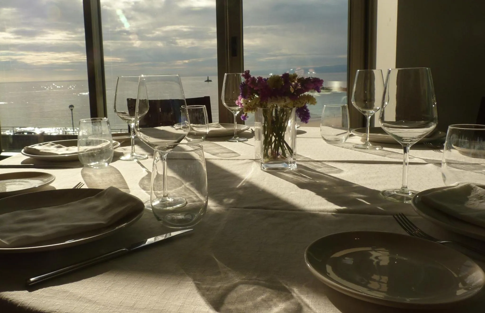 Une table dressée avec des couverts et des verres devant une grande baie vitrée donnant sur la mer.