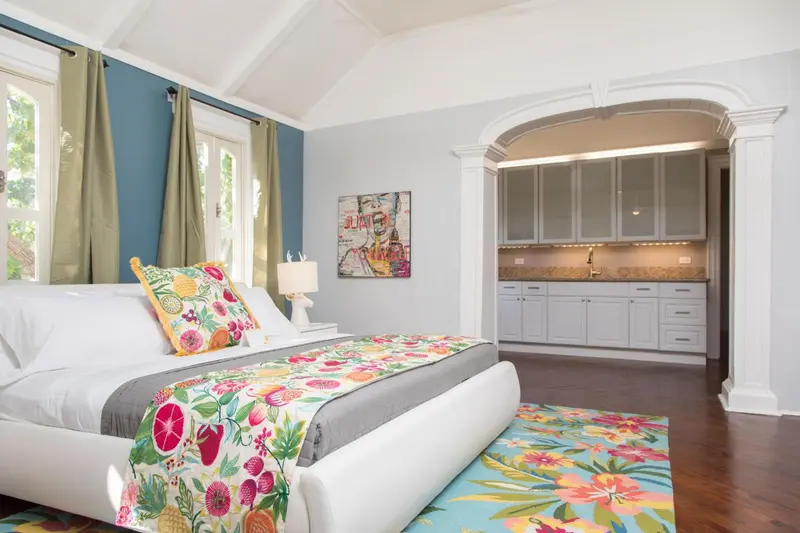 Une chambre lumineuse avec un grand lit décoré de motifs floraux et une kitchenette en fond.