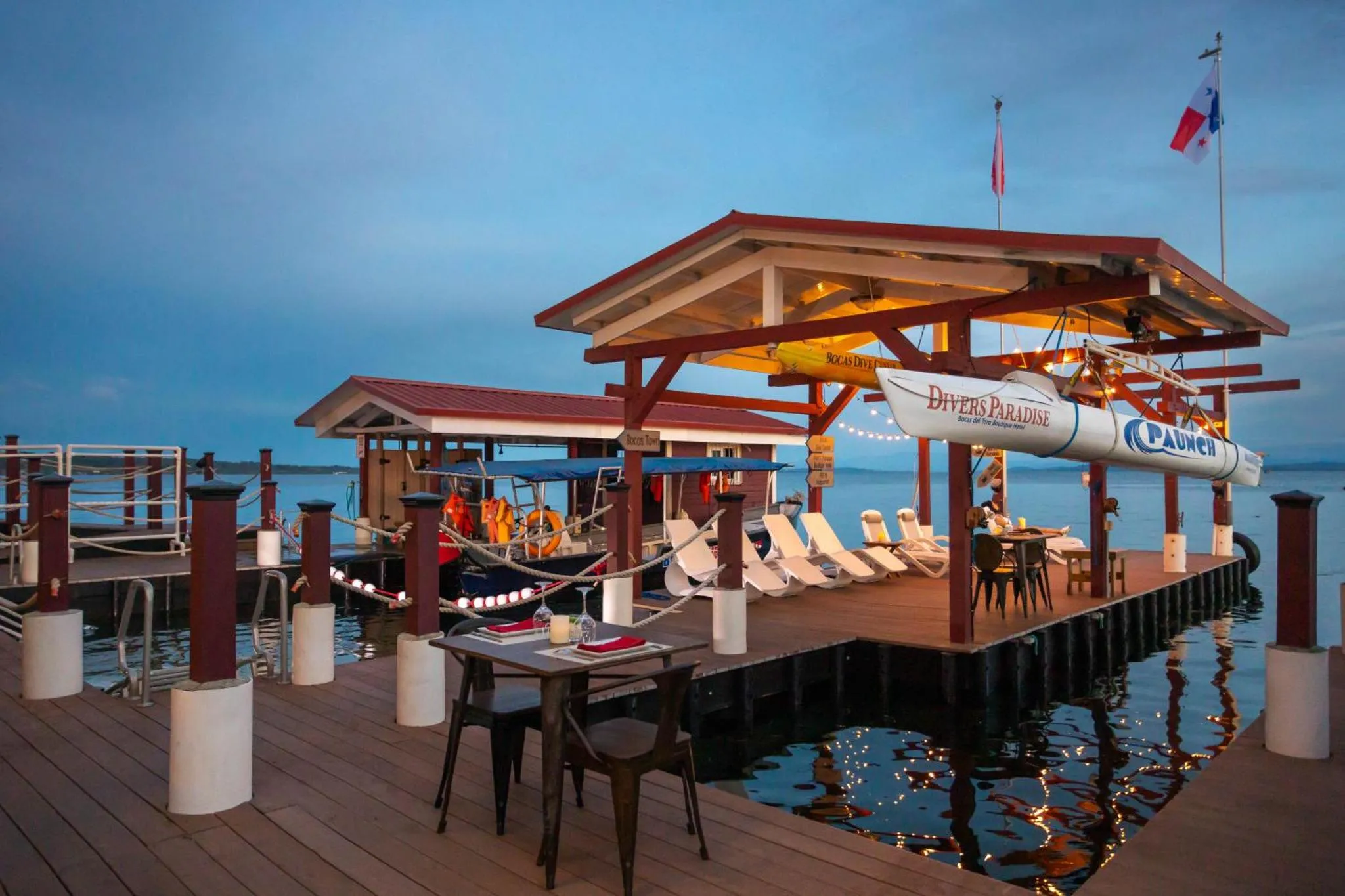 Ponton en bois avec tables, chaises et un abri illuminé au crépuscule.