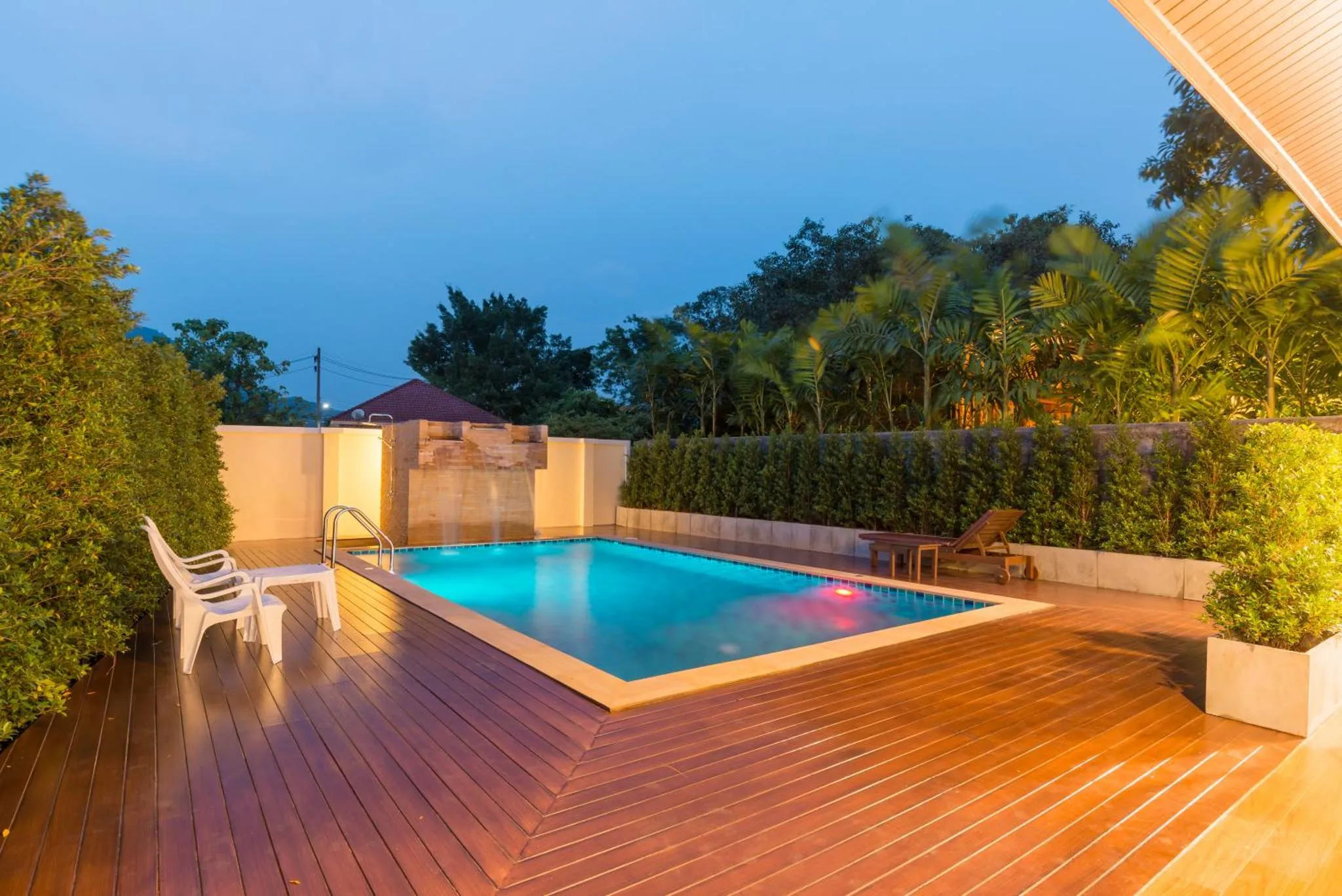 Une piscine rectangulaire illuminée au crépuscule, entourée d'une terrasse en bois et de végétation luxuriante.