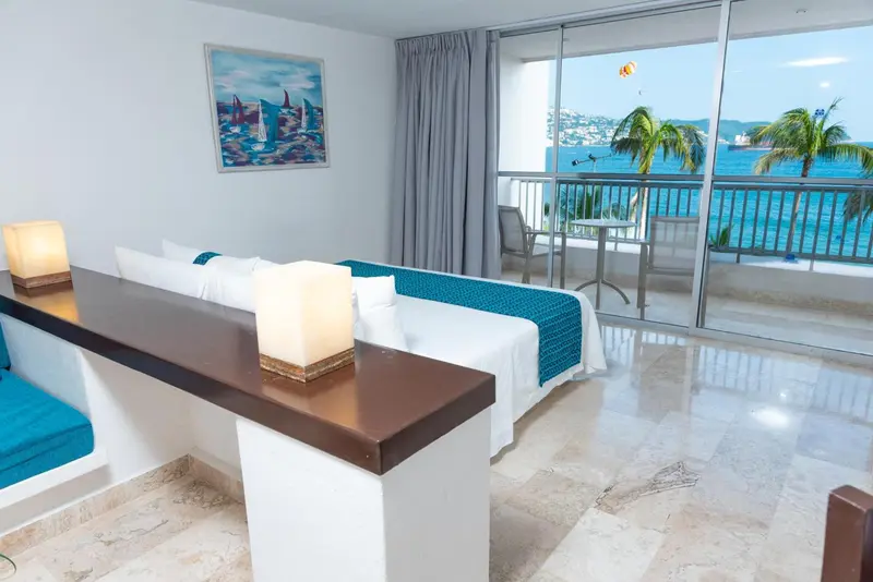 Chambre lumineuse avec un lit turquoise et une vue sur la mer depuis un balcon.