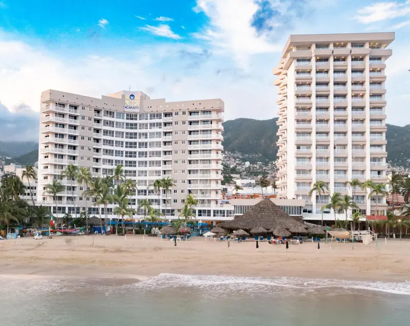 Vue d'un bâtiment blanc avec des balcons, bordant une plage de sable fin et une mer turquoise.