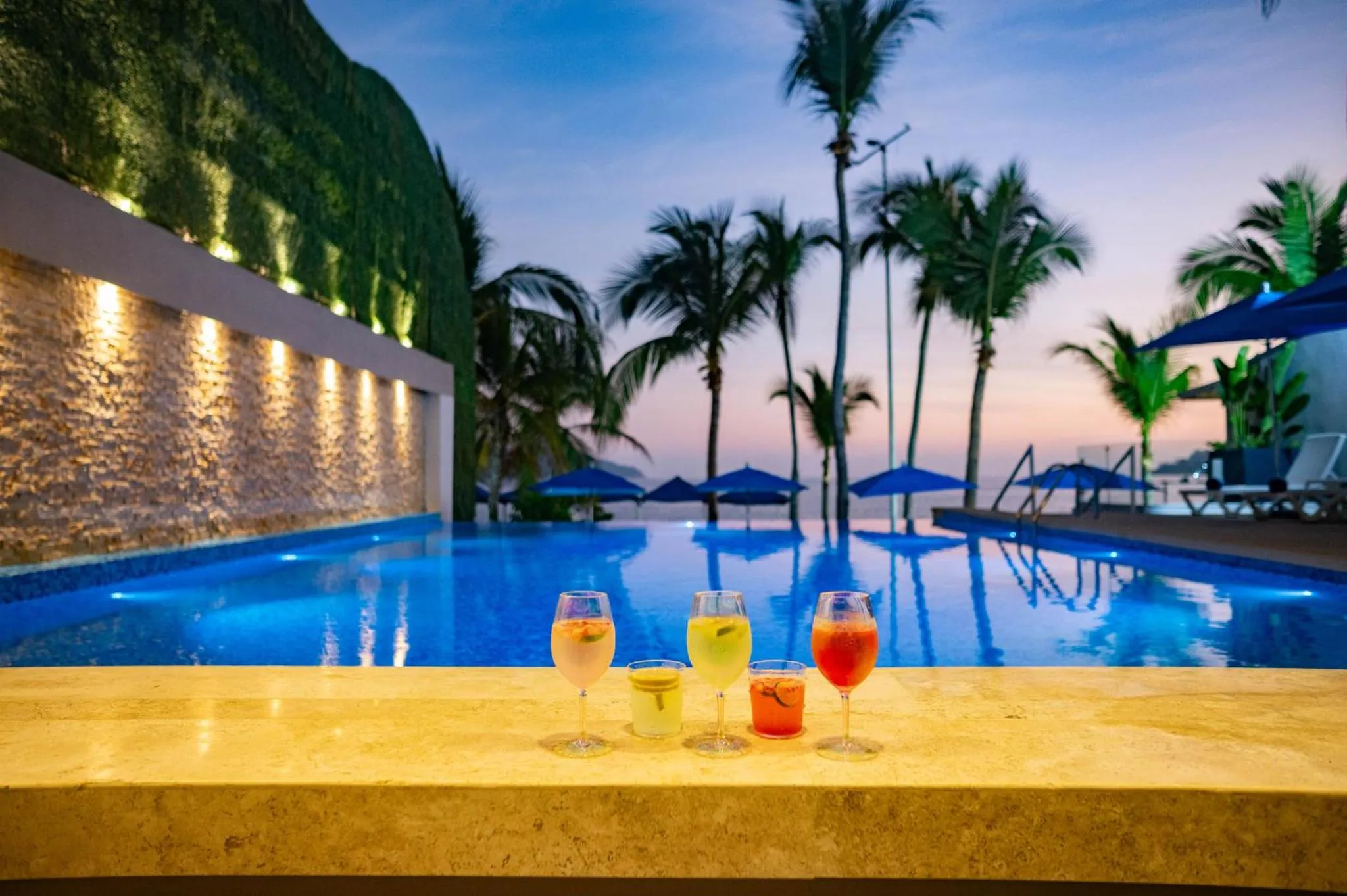 Des cocktails colorés sont posés sur le bord d'une piscine illuminée au crépuscule.