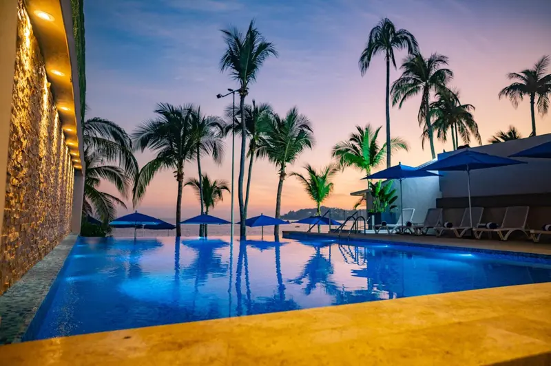 Piscine illuminée avec transats et palmiers au coucher du soleil.