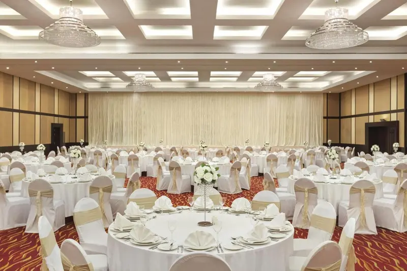 Une grande salle de réception est mise en scène avec des tables rondes nappées et décorées de fleurs.
