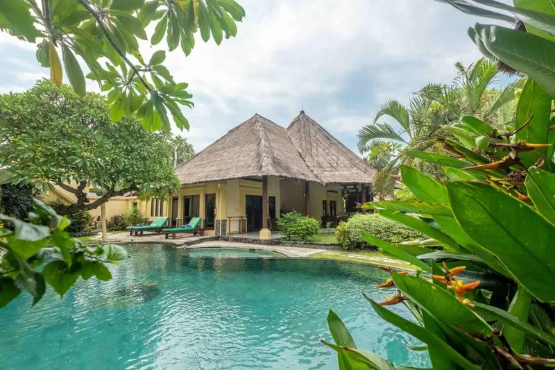 Bâtiment à toit de chaume près d'une piscine turquoise entourée de végétation tropicale.
