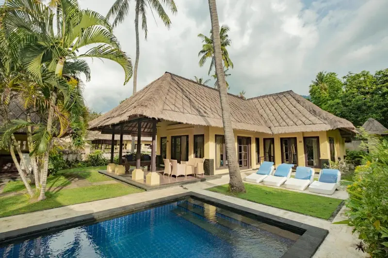 Bâtiment à toit de chaume avec transats et piscine devant un jardin tropical.