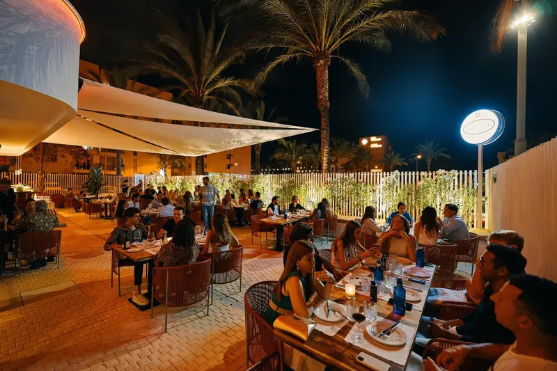 Une terrasse de restaurant animée avec des tables éclairées et des palmiers en arrière-plan.