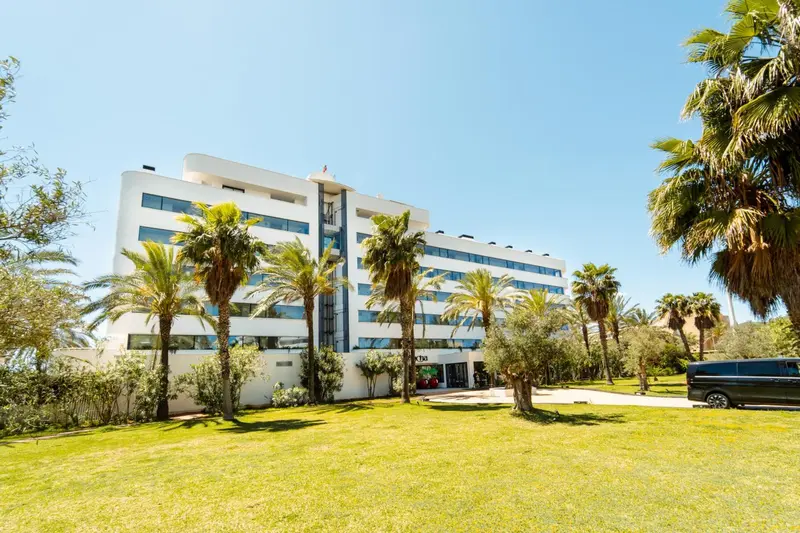 Bâtiment blanc moderne entouré de végétation luxuriante et de palmiers sous un ciel bleu.
