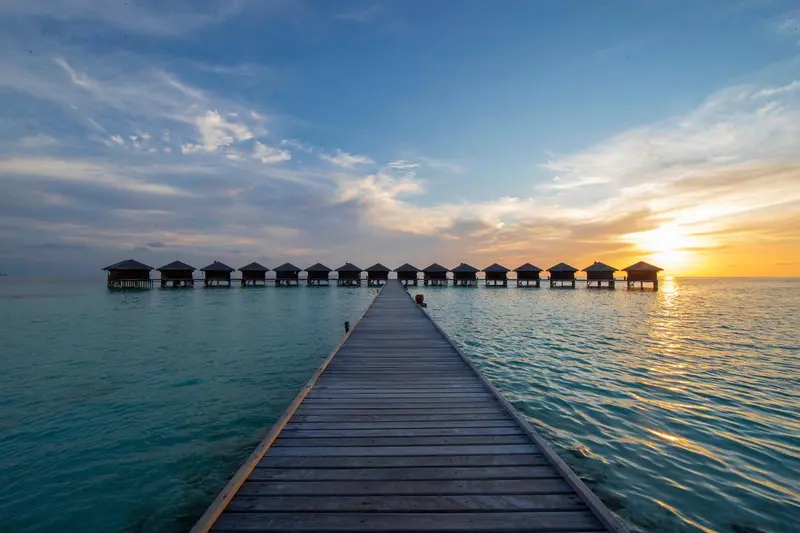 Un ponton en bois mène à des bungalows sur pilotis au coucher du soleil.
