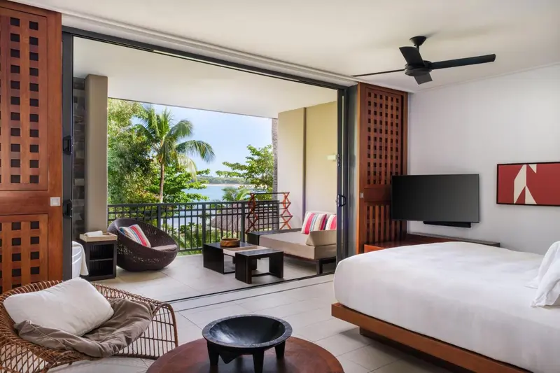 Une chambre lumineuse s'ouvre sur une terrasse avec vue sur la végétation et la mer.