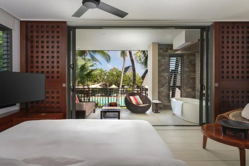 Une chambre lumineuse s'ouvre sur une terrasse avec vue sur une piscine et une végétation tropicale.