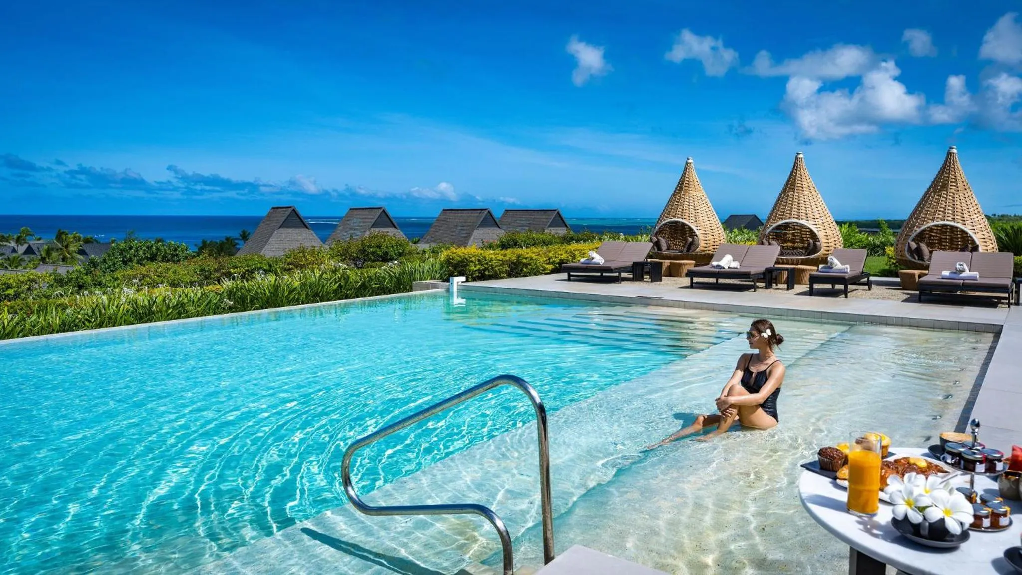 Une femme se détend au bord d'une piscine avec une vue sur la mer et des bungalows.