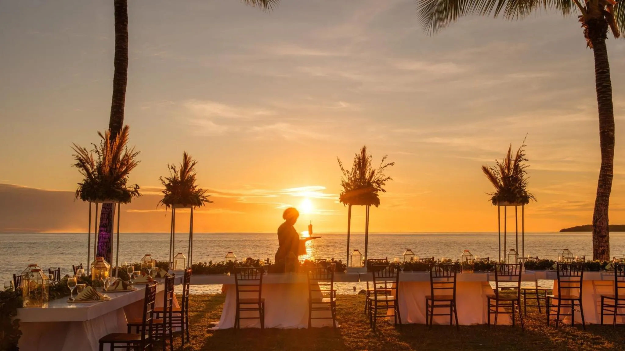 Une personne sert des boissons sur une terrasse face à la mer au coucher du soleil.