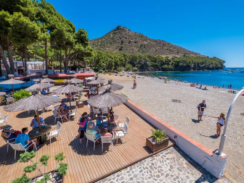 Terrasse de restaurant en bois avec parasols et tables, donnant sur une plage de sable et la mer.