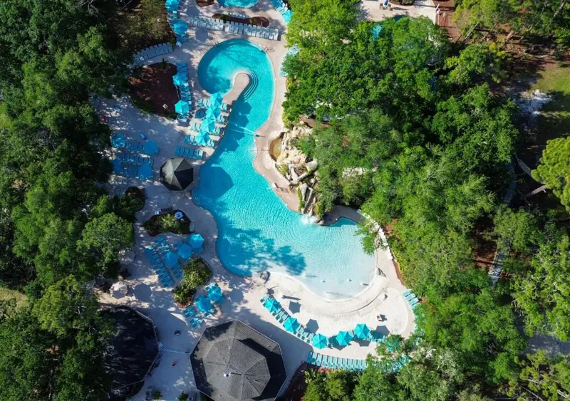 Vue aérienne d'une piscine sinueuse entourée de végétation luxuriante et de transats bleus.