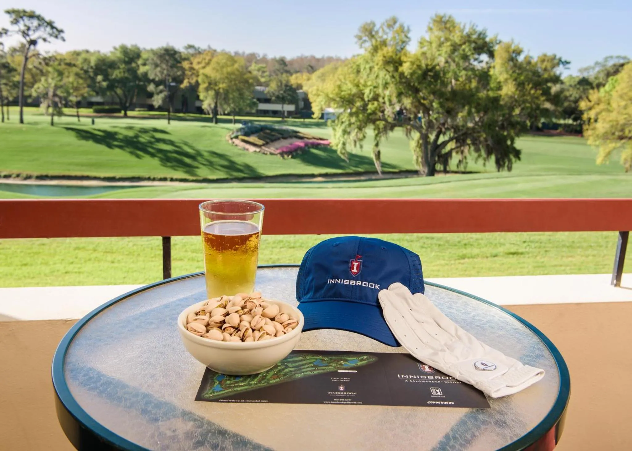 Un verre de bière, des cacahuètes et une casquette reposent sur une table d'extérieur avec vue sur un terrain de golf verdoyant.
