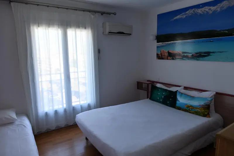 Chambre lumineuse avec un lit blanc et une vue sur la mer à travers une fenêtre.