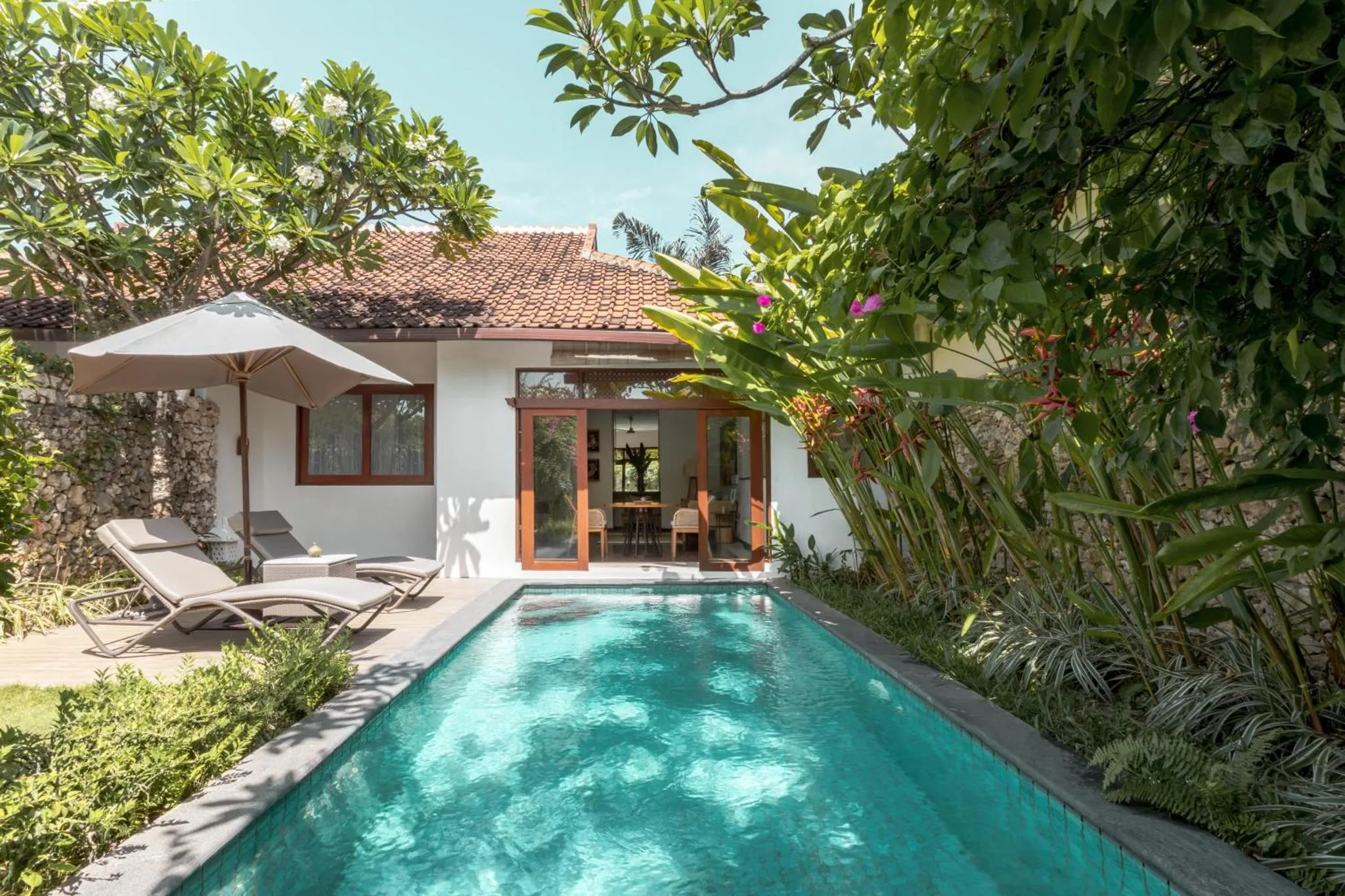 Petite piscine rectangulaire devant une maison blanche avec une terrasse et des transats.