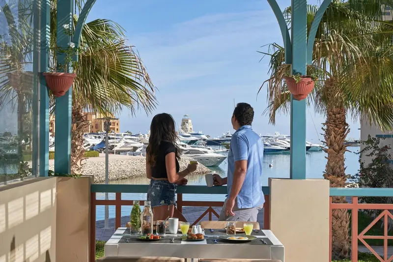 Deux personnes admirent la vue sur une marina ensoleillée depuis une terrasse.
