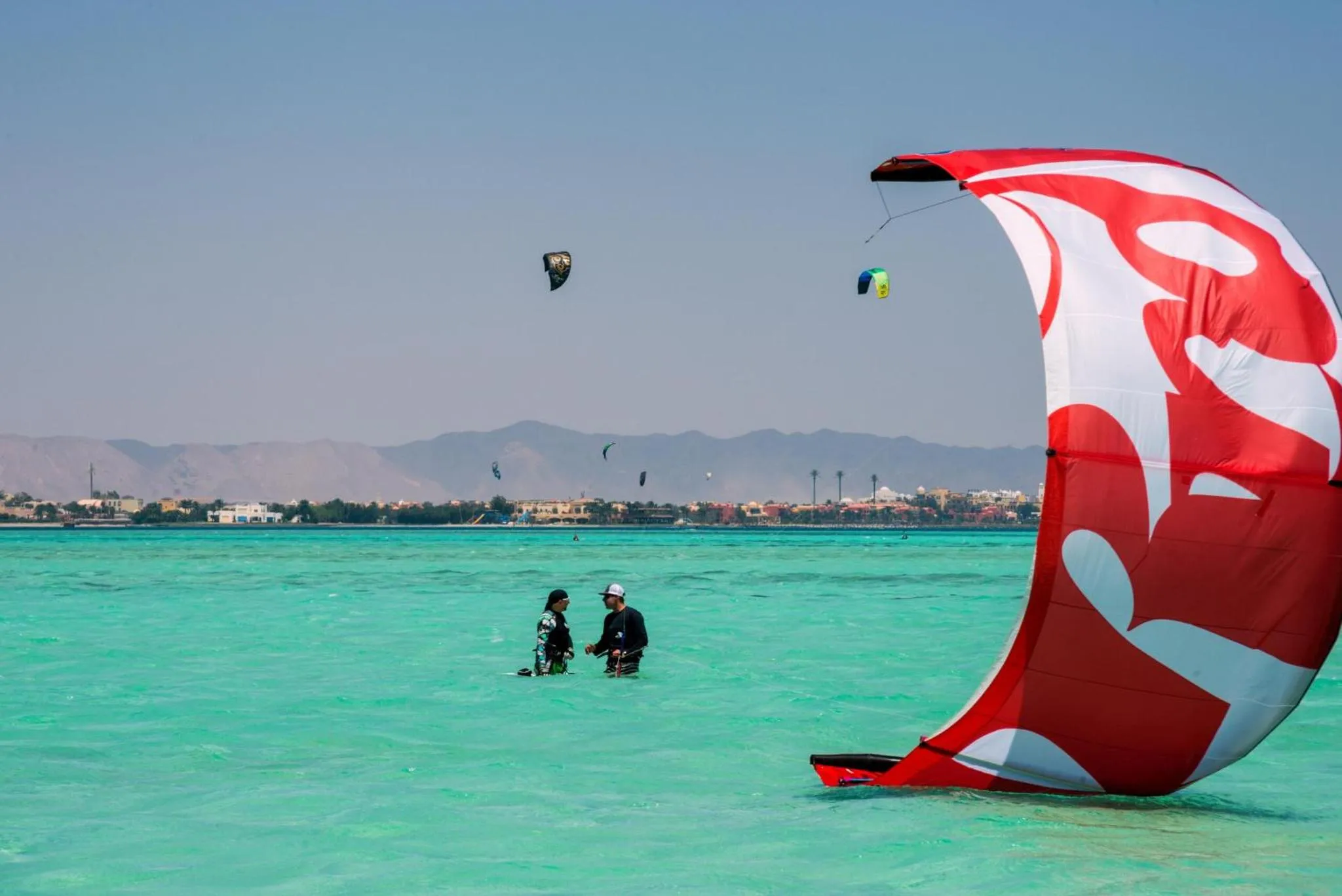 Deux personnes se tiennent dans une eau turquoise peu profonde avec des cerfs-volants colorés dans le ciel.