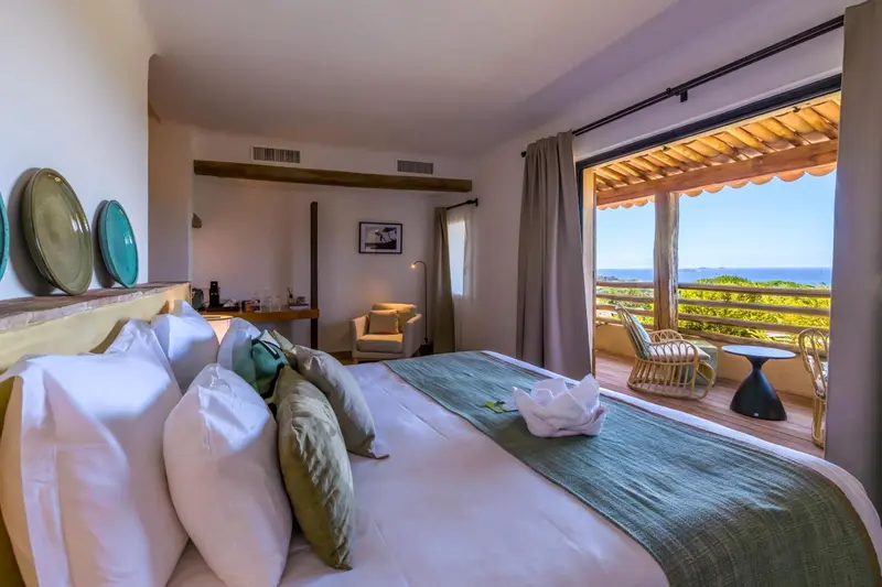 Chambre lumineuse avec un grand lit et une vue sur la mer depuis une terrasse.