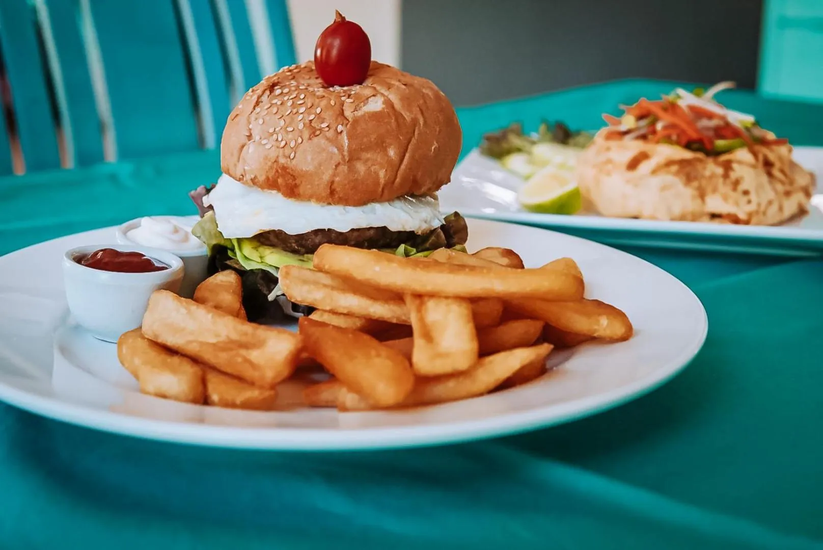 Assiette de hamburger, frites et salade colorée sur une table turquoise.