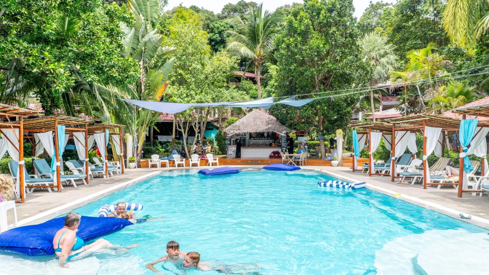 Une piscine bordée de transats sous des structures ombragées, avec des personnes se baignant.