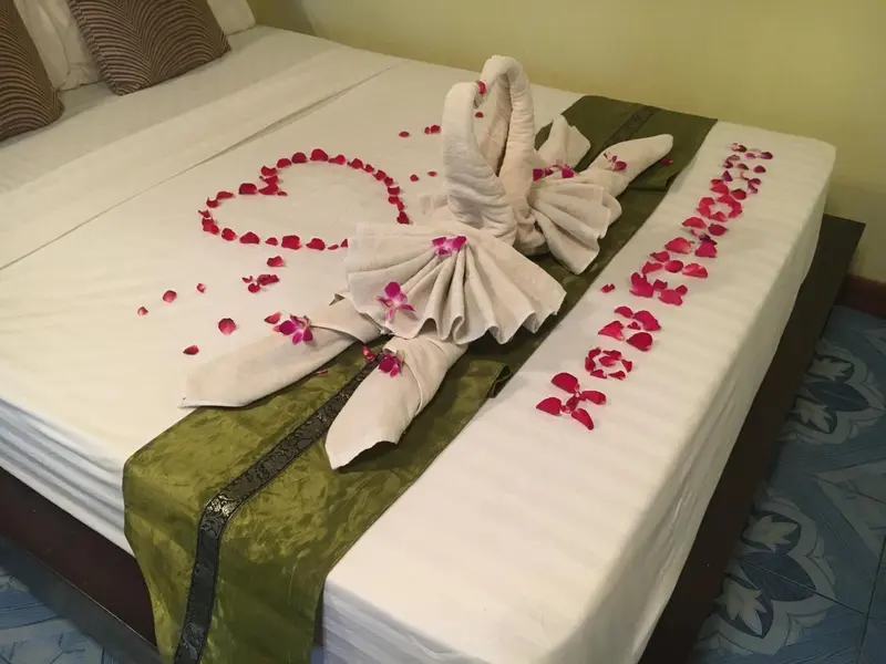Un lit recouvert de draps blancs est décoré de serviettes en forme de cygnes et de pétales de fleurs roses.