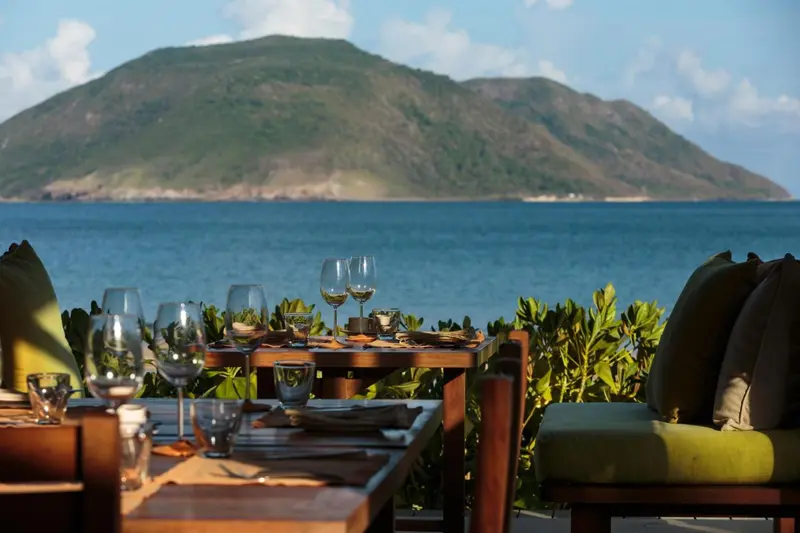 Une table est dressée avec des verres à vin et de la vaisselle face à la mer et à une colline verdoyante.