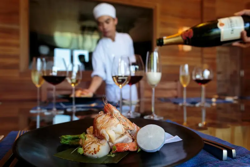 Une assiette de fruits de mer est présentée au premier plan, tandis qu'un serveur verse du vin en arrière-plan.