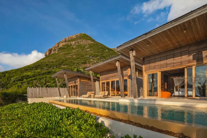 Une piscine turquoise borde des logements en bois avec une colline verdoyante en arrière-plan.