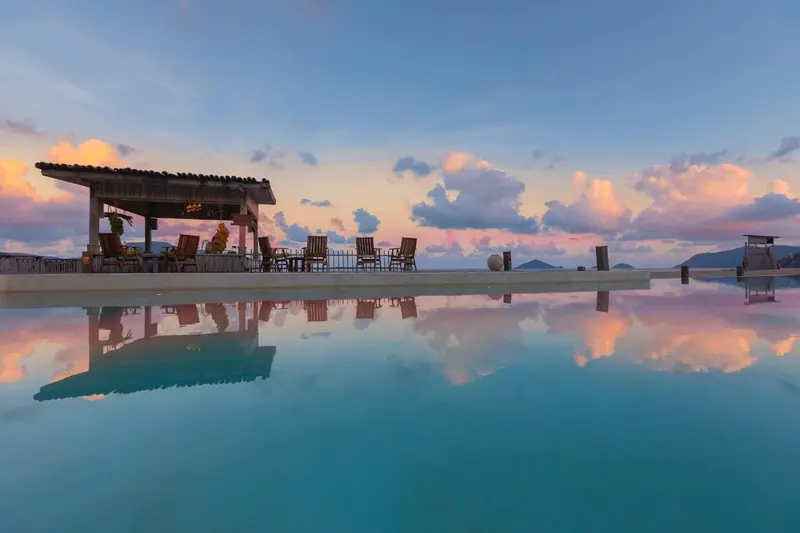Une piscine à débordement reflète le ciel crépusculaire et un espace détente ombragé.